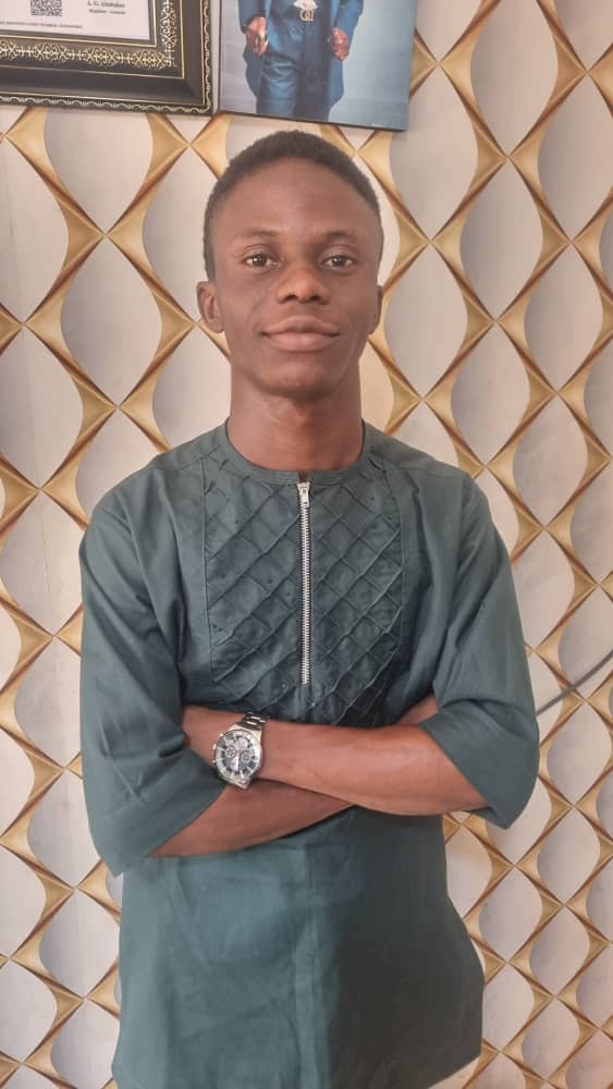Young man in a green zippered shirt posing with arms crossed against geometric patterned wallpaper.